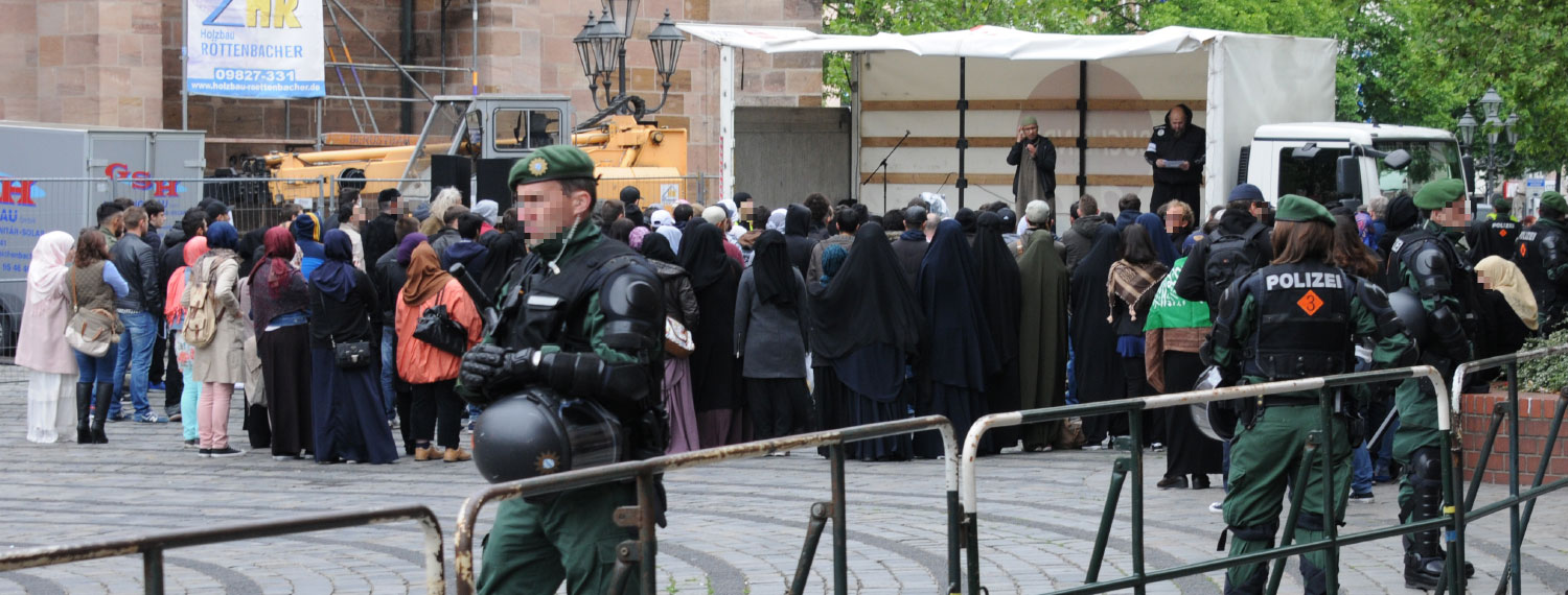 Menschengruppe vor einer Bühne. Polizisten bewachenden den Bereich.